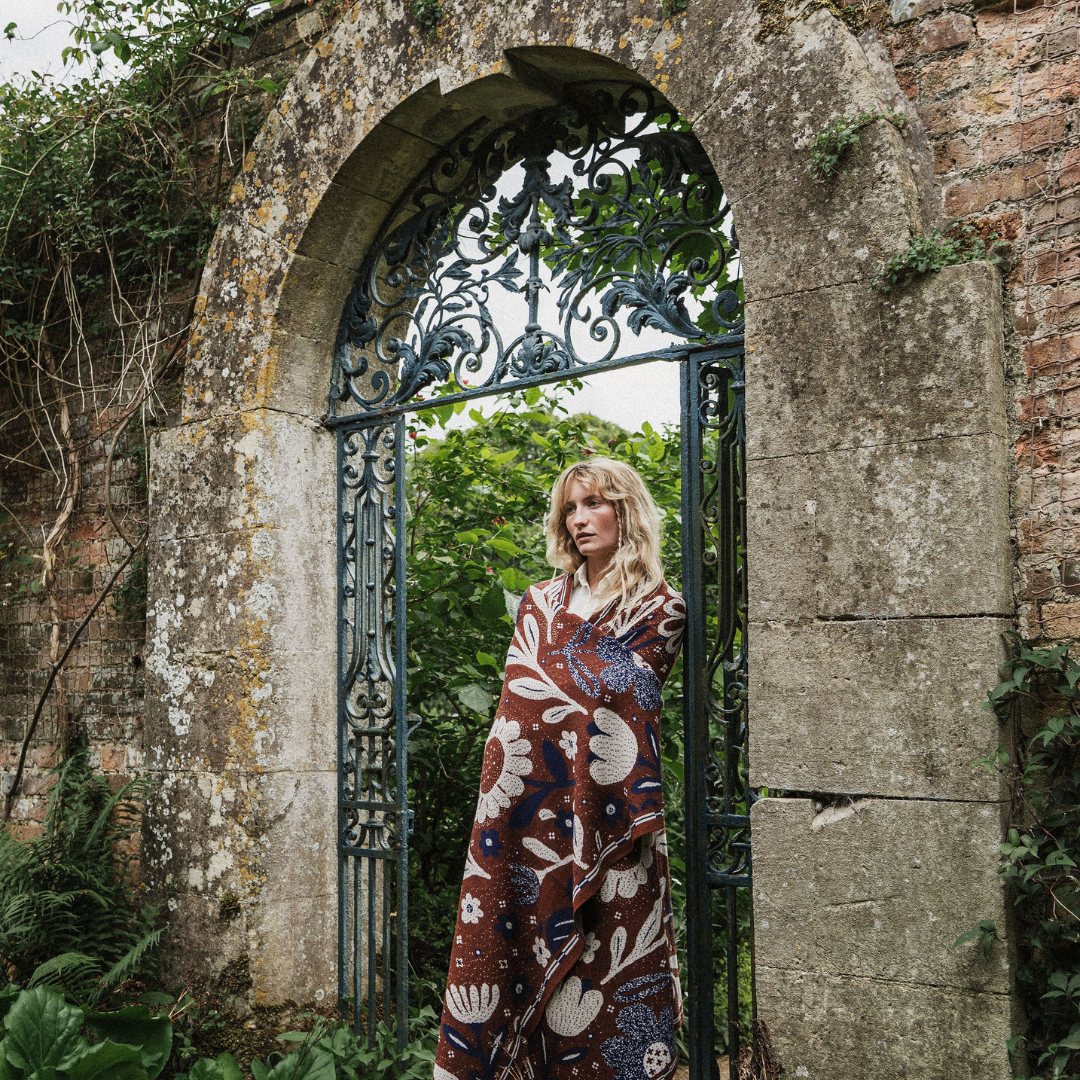 A woman stands in an arched stone gate surrounded by greenery, wrapped in a long patterned blanket or throw. The fabric has a brown base with large cream and blue floral motifs.