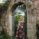 A woman stands in an arched stone gate surrounded by greenery, wrapped in a long patterned blanket or throw. The fabric has a brown base with large cream and blue floral motifs.