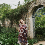 A woman stands in a garden setting wrapped in a patterned blanket. The blanket features a dark red base with cream, blue, and brown floral and abstract botanical patterns.