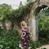 A woman stands in a garden setting wrapped in a patterned blanket. The blanket features a dark red base with cream, blue, and brown floral and abstract botanical patterns.