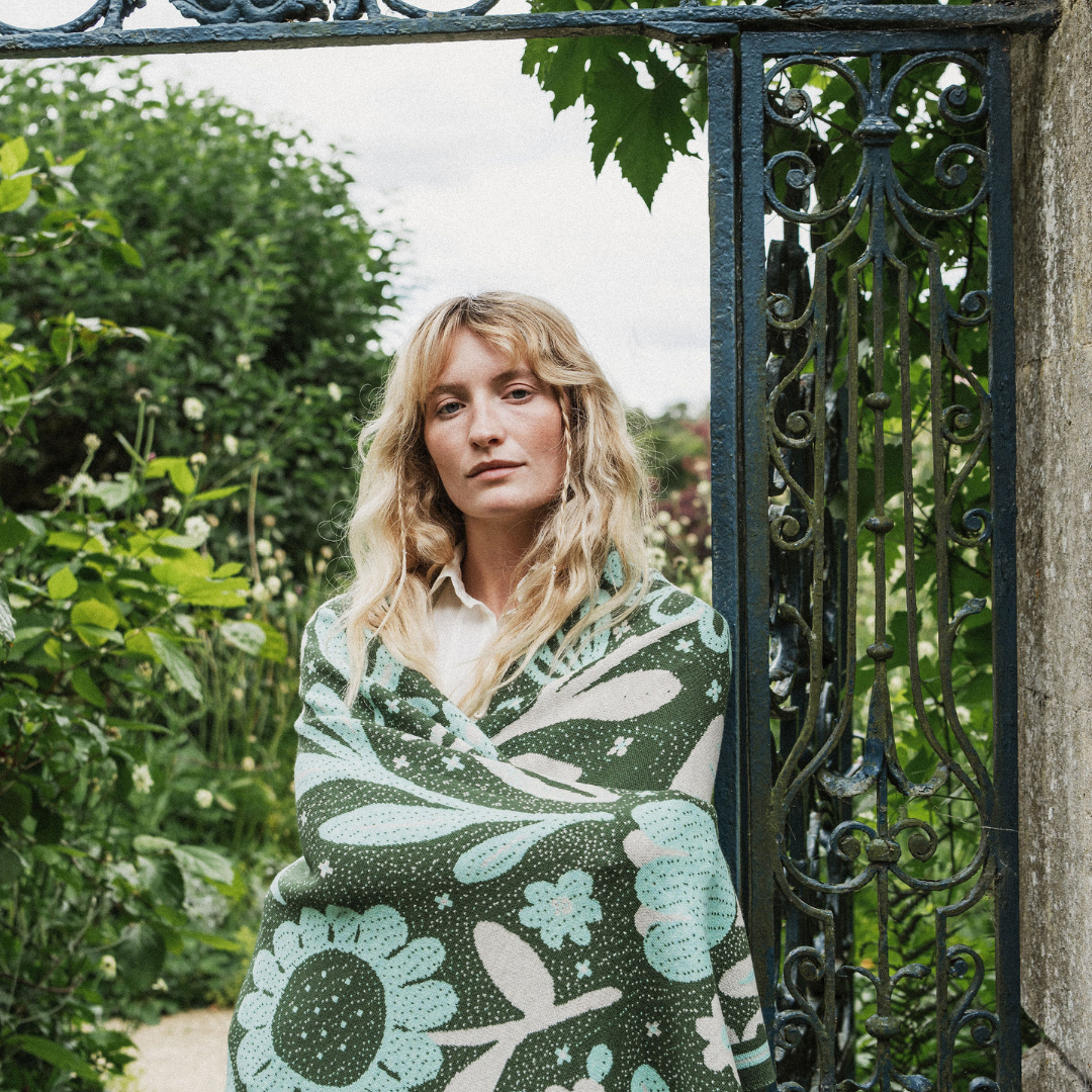 A woman with blonde hair stands behind an ornate black metal garden gate, wrapped in a dark green blanket featuring a large light blue and cream floral pattern.