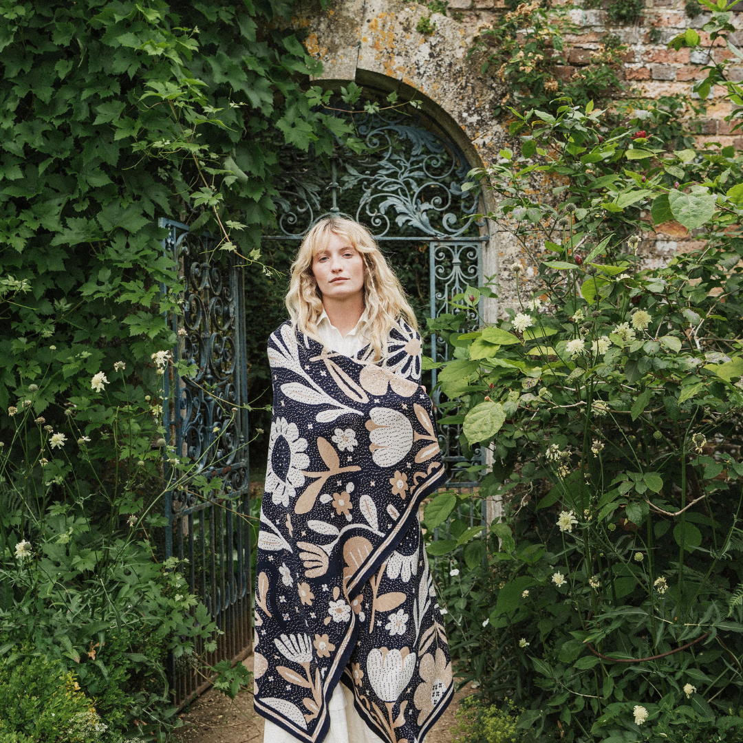A woman stands in a garden courtyard in front of an arched iron gate, wrapped in a dark navy knit blanket with a beige and cream floral pattern.