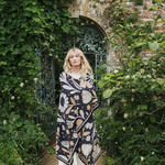 A woman stands in a garden courtyard in front of an arched iron gate, wrapped in a dark navy knit blanket with a beige and cream floral pattern.