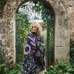 A woman wrapped in a dark knit blanket with a large purple and green floral pattern, standing in front of an arched stone gate with green foliage.
