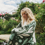 A woman wrapped in a dark green blanket with a light green and cream botanical pattern, standing in a lush garden.