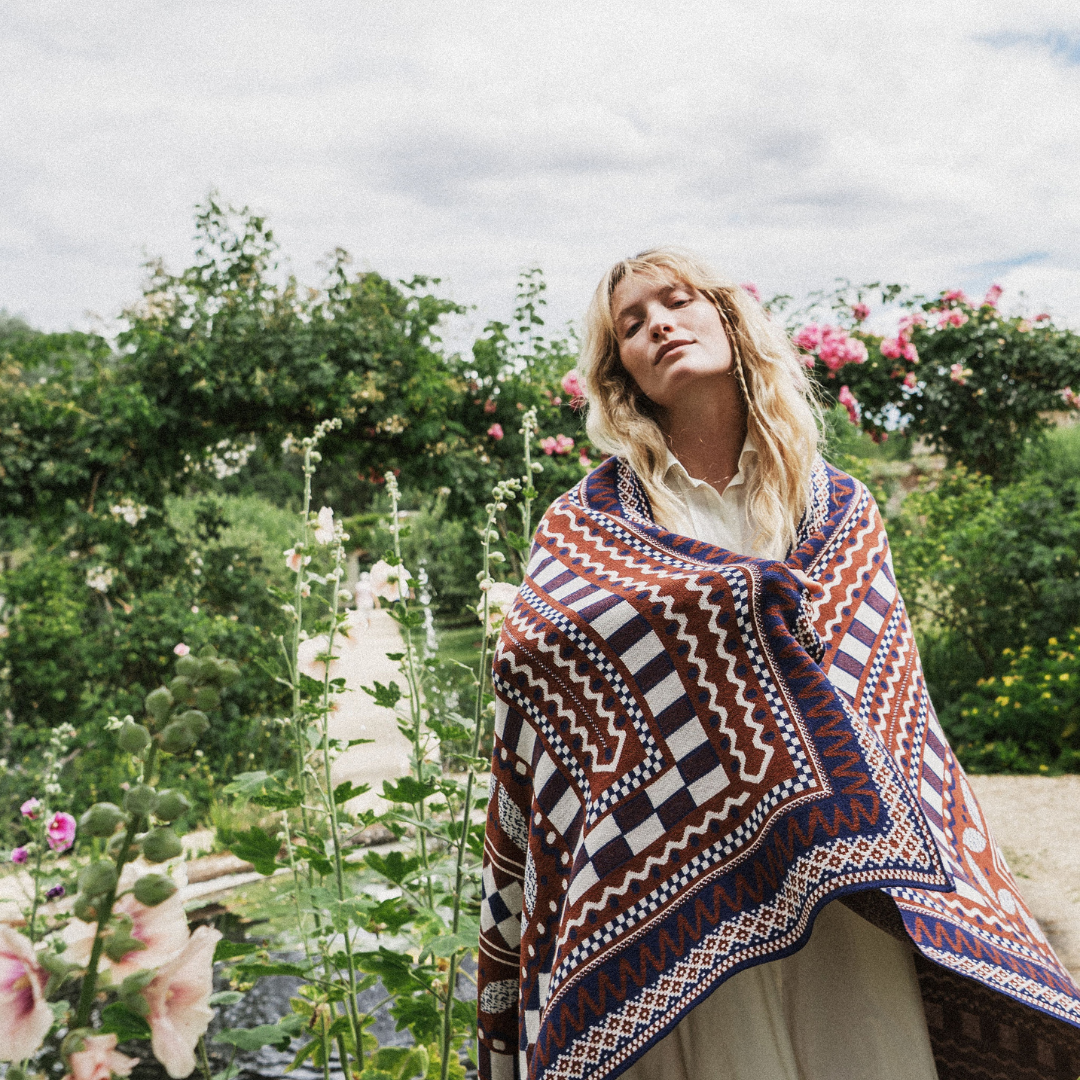 A woman outdoors in a lush garden wearing a large patterned knit shawl over a cream dress. The shawl features a geometric tribal-style pattern in rust red, navy blue, white, and brown.
