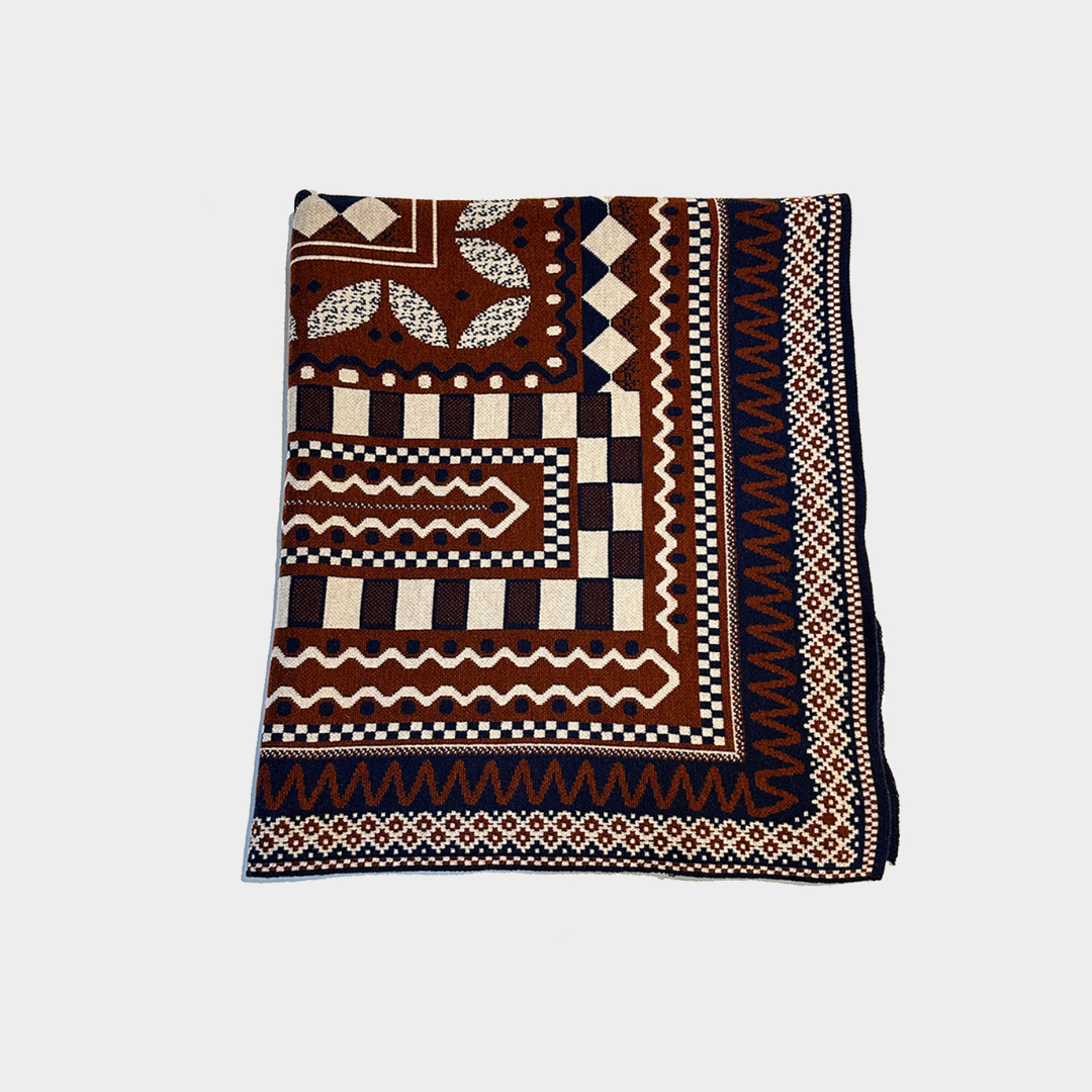 A folded square blanket with an intricate geometric and tribal-inspired pattern in dark brown, black, cream, and navy blue, displayed flat against a light gray background.