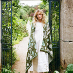 A woman stands in an open black wrought-iron garden gate, wearing a white dress and a large green patterned blanket with pale blue and white botanical motifs. The background shows a lush green garden path and stone wall.