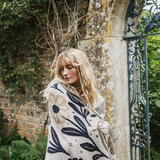 A woman with long blonde hair wrapped in a cream knit blanket featuring navy blue and beige botanical patterns, standing outdoors next to a stone wall and an ornate dark metal gate.