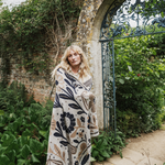 A woman stands in a garden courtyard wrapped in a cream-colored knit blanket featuring a navy blue and tan botanical pattern. She is positioned in front of a stone archway and an open dark green wrought iron gate, surrounded by lush green plants.