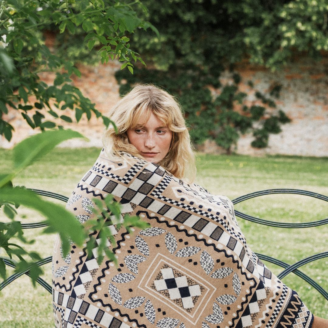 A woman sits on a black metal garden bench wrapped in a beige bohemian patterned blanket, with green foliage and a stone wall in the background.