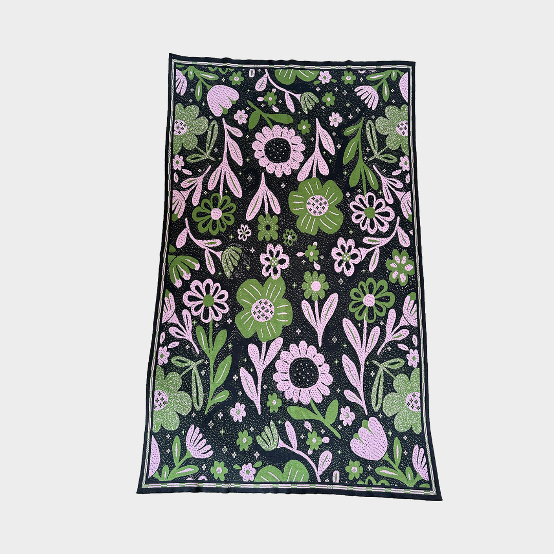 A knitted blanket with a dense floral pattern on a black background, featuring pink and green flowers, leaves, and stems, bordered by a thin dark edge, shown flat against a light gray background.