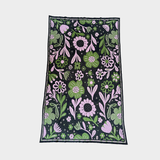 A knitted blanket with a dense floral pattern on a black background, featuring pink and green flowers, leaves, and stems, bordered by a thin dark edge, shown flat against a light gray background.