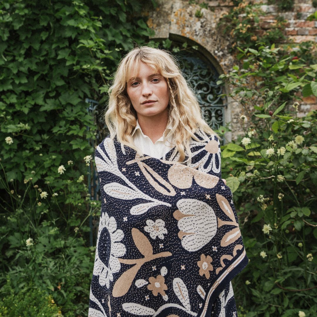 A woman with blonde hair stands outdoors in front of a stone wall covered in green ivy, wearing a navy blue blanket with a large-scale cream and beige floral pattern over a white collared shirt.