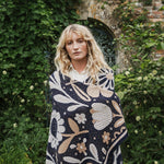 A woman with blonde hair stands outdoors in front of a stone wall covered in green ivy, wearing a navy blue blanket with a large-scale cream and beige floral pattern over a white collared shirt.
