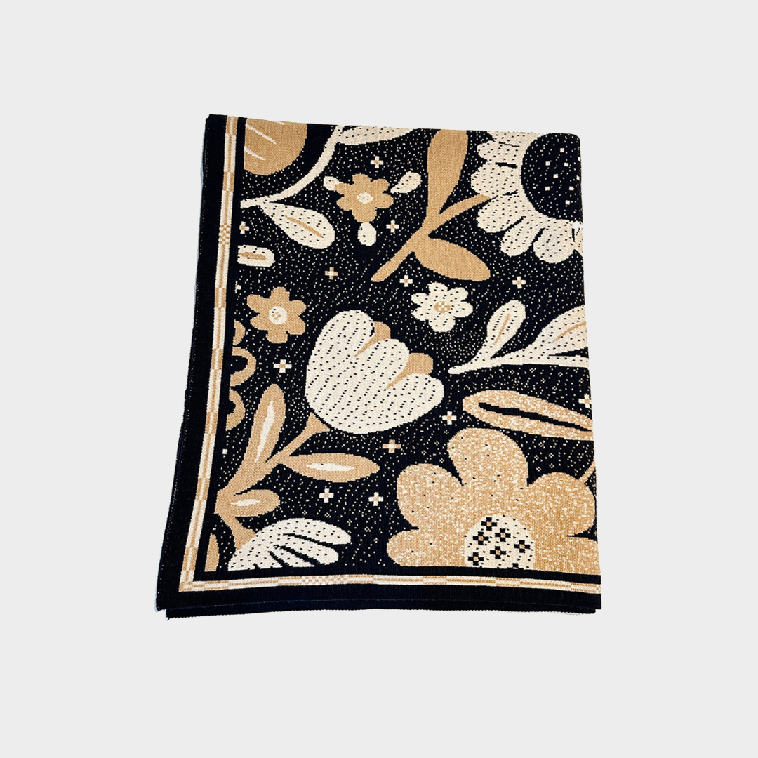 A folded square blanket in a floral pattern in beige, cream, and black, displayed flat against a light gray background.