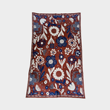 A rectangular blanket with a dense floral and botanical pattern, featuring white and navy blue motifs on a rust red background, photographed flat against a light gray background.