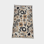 A rectangular blanket with an all-over floral pattern, shown flat against a light gray background. The design features black, dark brown, and tan flowers and leaves on a warm off-white base.