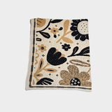 A folded woven textile blanket, featuring a bohemian-style floral pattern in black, tan, and beige on a cream background, with a striped border.