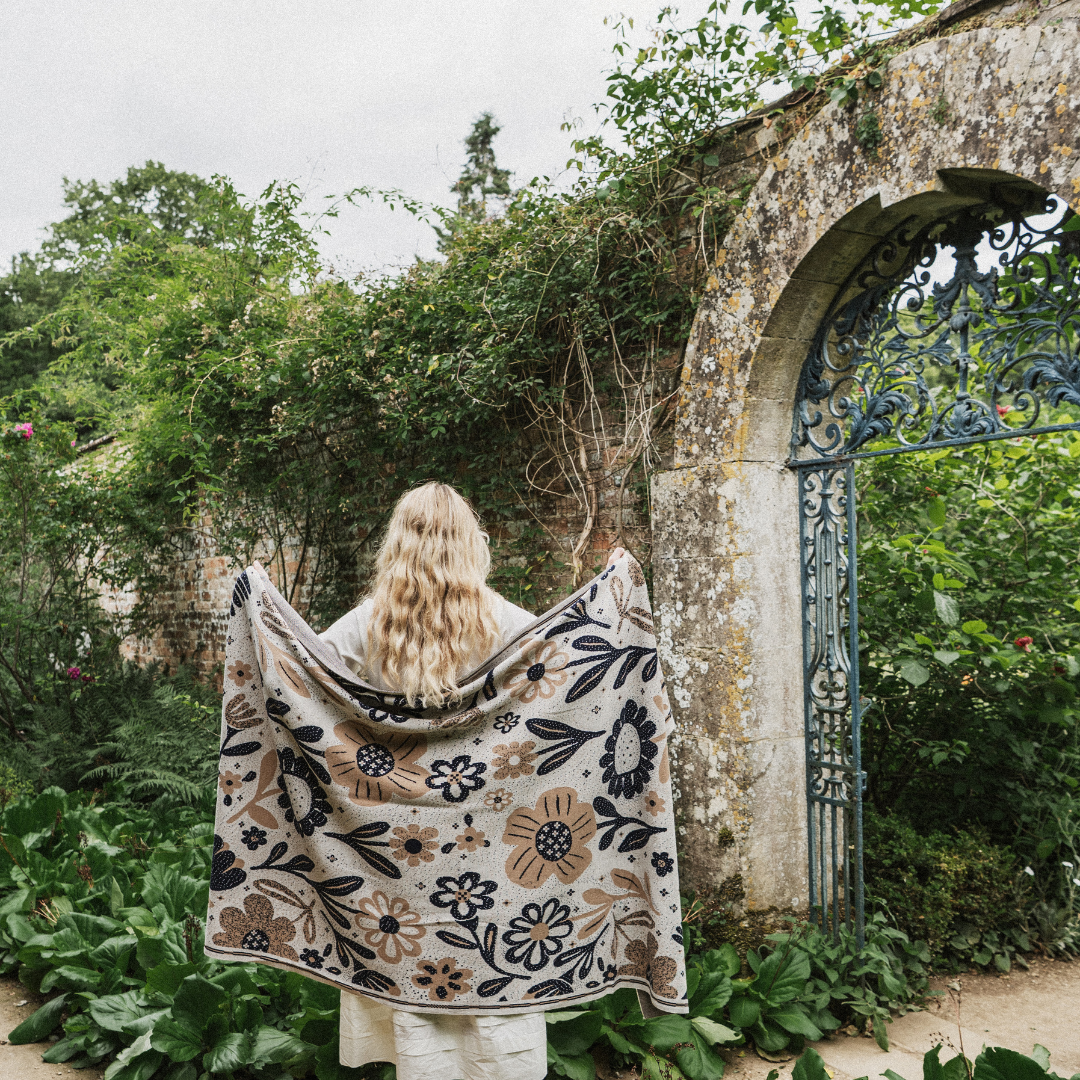 A woman stands with her back to the camera in a garden courtyard, holding up a large cream-colored floral blanket or throw with black and brown botanical patterns. She is positioned in front of a weathered stone archway and lush green foliage.