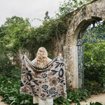 A woman stands with her back to the camera in a garden courtyard, holding up a large cream-colored floral blanket or throw with black and brown botanical patterns. She is positioned in front of a weathered stone archway and lush green foliage.