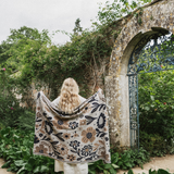 A woman stands with her back to the camera in a garden courtyard, holding up a large cream-colored floral blanket or throw with black and brown botanical patterns. She is positioned in front of a weathered stone archway and lush green foliage.