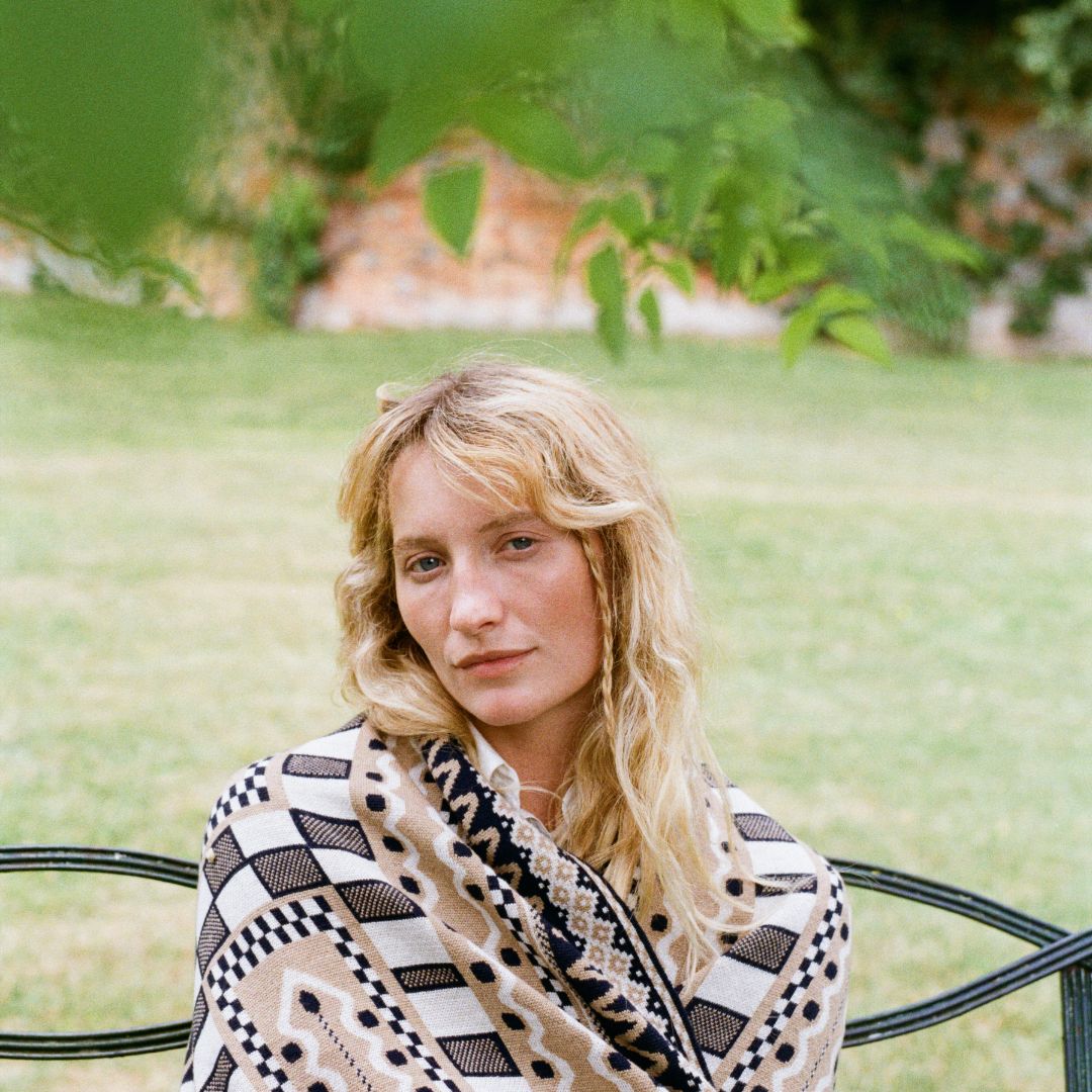 A woman with blonde hair sits on a black metal bench in a grassy outdoor garden, wearing a beige, black, and white patterned knit blanket.