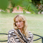 A woman with blonde hair sits on a black metal bench in a grassy outdoor garden, wearing a beige, black, and white patterned knit blanket.
