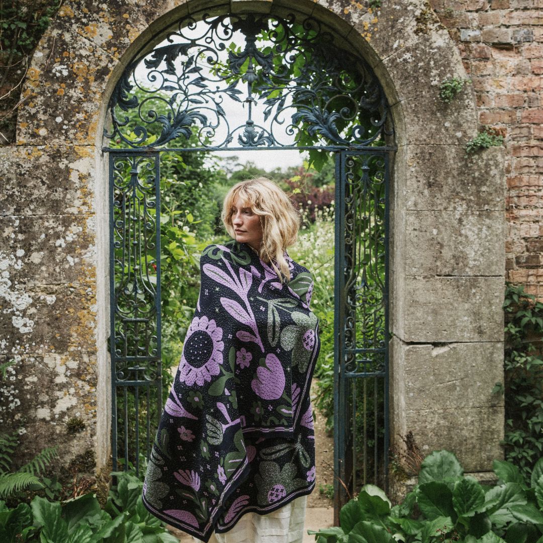 A woman stands in front of an arched stone gate with greenery and brickwork, wrapped in a large black throw blanket featuring a purple and green floral pattern.
