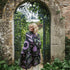 A woman stands in front of an arched stone gate with greenery and brickwork, wrapped in a large black throw blanket featuring a purple and green floral pattern.