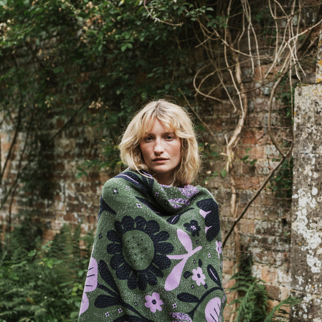 A woman with blonde hair stands outdoors in front of a rustic brick wall covered in greenery, wrapped in a dark green knit blanket featuring large black and lavender floral patterns.