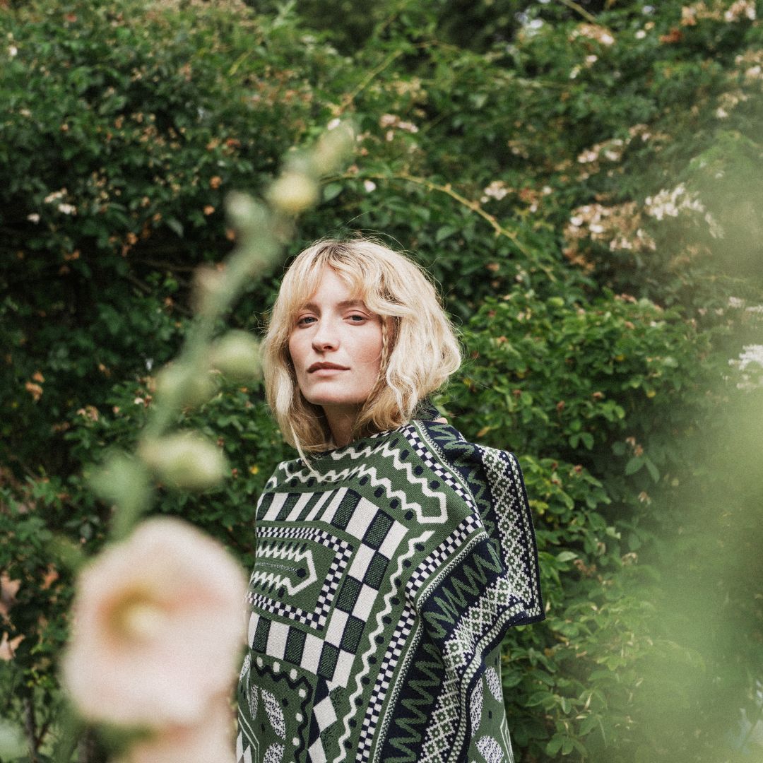 A woman with blonde hair stands outdoors in front of dense green foliage, wearing a dark green and black patterned blanket with geometric and tribal-inspired motifs.