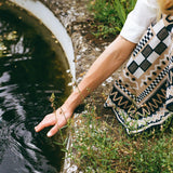 A person wearing a white short-sleeve top and a black, beige, and white geometric patterned blanket leans over the edge of a pond, touching the dark water with their hand.
