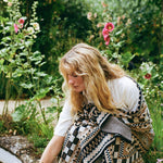 A woman with long blonde hair is crouching in a lush garden, wrapped in a patterned knit blanket or throw with black, beige, and white geometric designs.