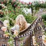 A woman with long blonde hair sits outdoors in a garden, viewed from behind, wrapped in a large patterned throw blanket. The blanket features a bohemian-style geometric design in beige, black, and white.