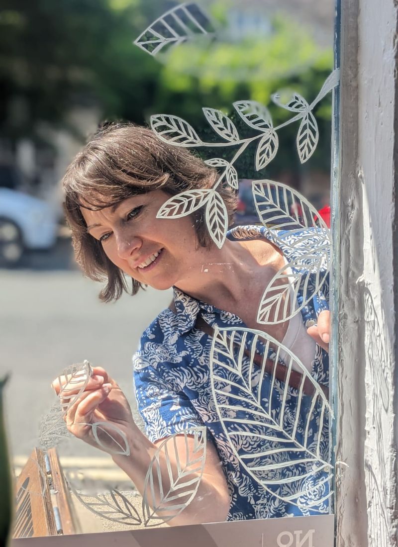 A woman with short brown hair smiles while applying a white leaf-patterned decal to a glass window. She is wearing a blue and white patterned blouse, and the outdoor street scene is softly blurred in the background.