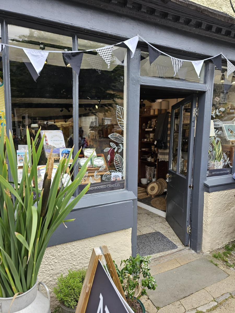 A storefront with dark gray painted trim and open windows, showing a shop interior with shelves and decor. A string of black-and-white bunting flags hangs across the top, and potted plants sit in the foreground on a stone sidewalk.
