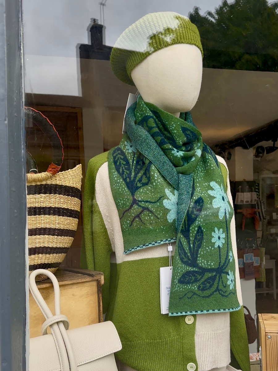 A green knit scarf with a floral and tree pattern in shades of blue and white, styled on a mannequin wearing a matching green beanie and cardigan in a shop window display.
