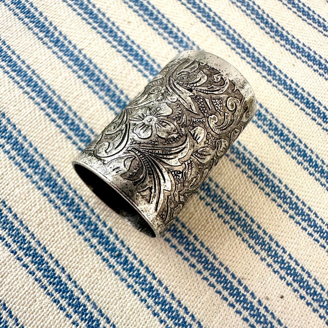 An ornate silver-toned metal scarf slider with intricate floral and scroll engravings, resting on a blue and cream striped woven fabric background.