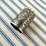 An ornate silver-toned metal scarf slider with intricate floral and scroll engravings, resting on a blue and cream striped woven fabric background.