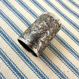 An ornate silver-toned metal scarf slider with intricate floral and scroll engravings, resting on a blue and cream striped woven fabric background.