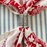 A close-up of a silver metal scarf slider with an embossed floral pattern, used as a napkin ring to hold a cream-colored scarf with a red floral print, set against a blue and white striped fabric background.