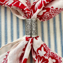 A close-up of a silver metal scarf slider with an embossed floral pattern, used as a napkin ring to hold a cream-colored scarf with a red floral print, set against a blue and white striped fabric background.