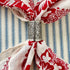 A close-up of a silver metal scarf slider with an embossed floral pattern, used as a napkin ring to hold a cream-colored scarf with a red floral print, set against a blue and white striped fabric background.