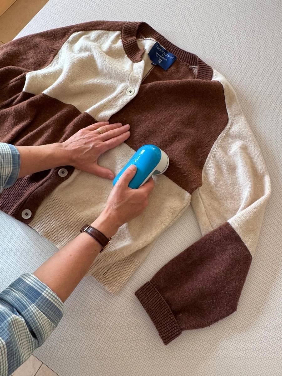 A person uses a blue handheld woll comb on a brown and cream color-blocked knit cardigan laid flat on a white surface.