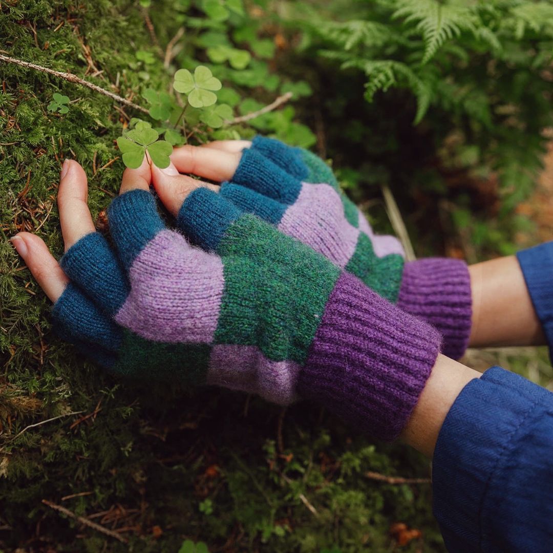 A close-up of a person's hands wearing chunky knit fingerless gloves with a patchwork pattern of teal, green, purple, and lavender, resting on mossy ground outdoors.