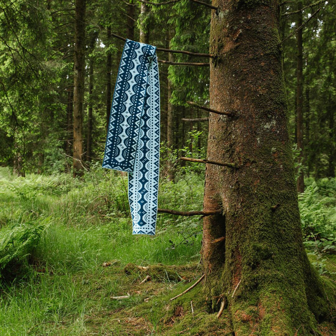 A blue and white patterned scarf, hangs from a rustic wooden rack attached to a mossy tree trunk in a green forest.
