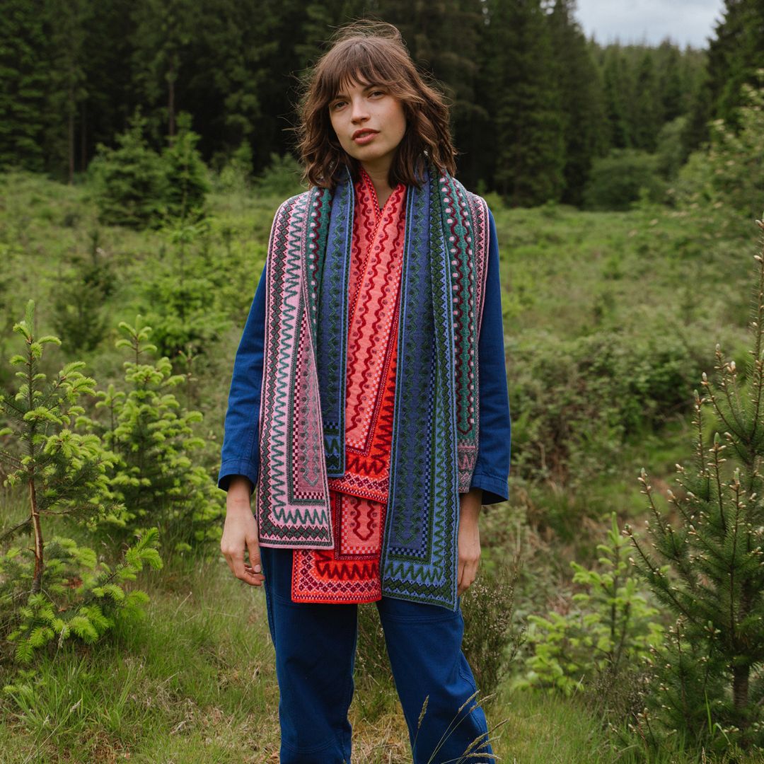 A woman standing outdoors in a grassy forest setting, wearing a long multicolor patterned knit scarf draped over a blue cardigan and blue pants.