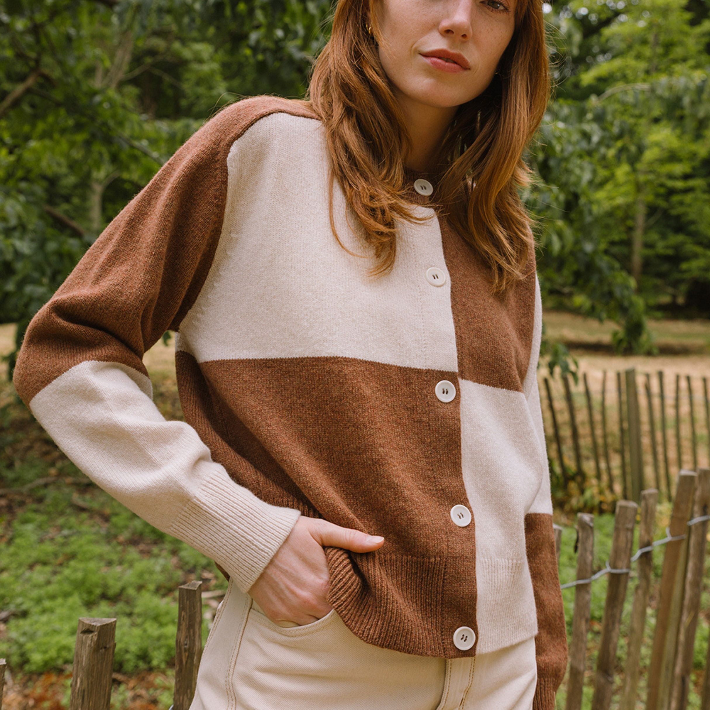 Woman wearing a brown and beige cardigan outdoors with greenery in the background