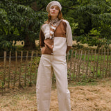Person wearing a brown and white cardigan with a scarf, standing in a field with trees in the background.